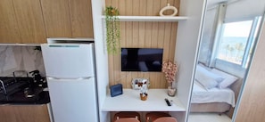 A compact kitchenette with a refrigerator, stove, and sink. A flat-screen TV is mounted on a wooden wall above a small table with a tablet, a flower vase, and a remote control. A bed with white bedding is visible through a window. - COPIA0108 - Edifício Essencial Piata (Salvador)