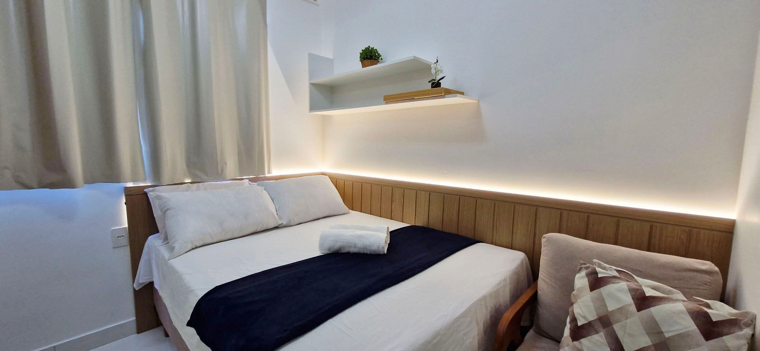 Quarto de hotel com cama, mesa de cabeceira, cadeira, prateleira na parede com uma planta e janela com cortinas.