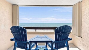 En balkong med två Adirondack-stolar och ett bord, med utsikt över en strand, havet och en klar himmel.
