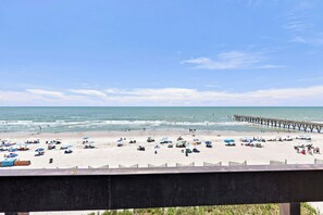 A beach with a pier extending into the water, multiple umbrellas, and people relaxing on the sand.