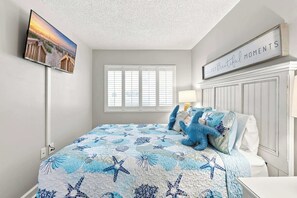 A bedroom with a bed, a television mounted on the wall, a window with blinds, and a sign above the bed. - Serenity at Sea Pointe Oceanfront Condo with Pool (North Myrtle Beach)