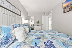 A bedroom with a bed, a bedside table, a dresser, a mirror, and a door. - Serenity at Sea Pointe Oceanfront Condo with Pool (North Myrtle Beach)