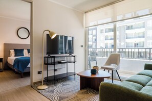 Ein modernes Hotelzimmer mit Bett, Fernseher auf einem Schrank, rundem Couchtisch, grünem Sofa und einem Balkon mit Stadtblick.