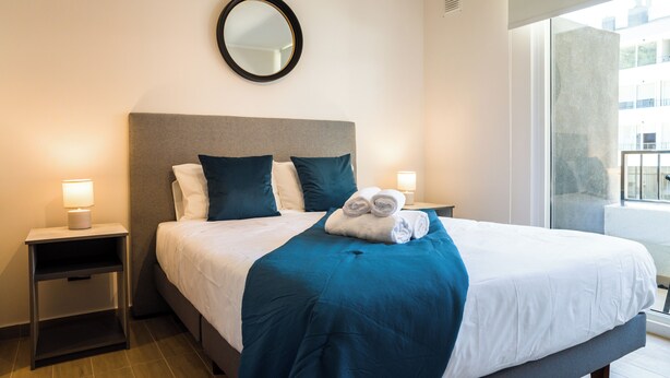 A hotel room with a bed, two bedside tables, a nightstand, a mirror, and a window with a view of buildings. - Inés Matte Urrejola Apartments by Andes STR (Providencia)