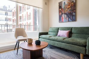 A modern living room with a green sofa, a beige armchair, a wooden coffee table, and a large window offering a view of a building with balconies. - Inés Matte Urrejola Apartments by Andes STR (Providencia)