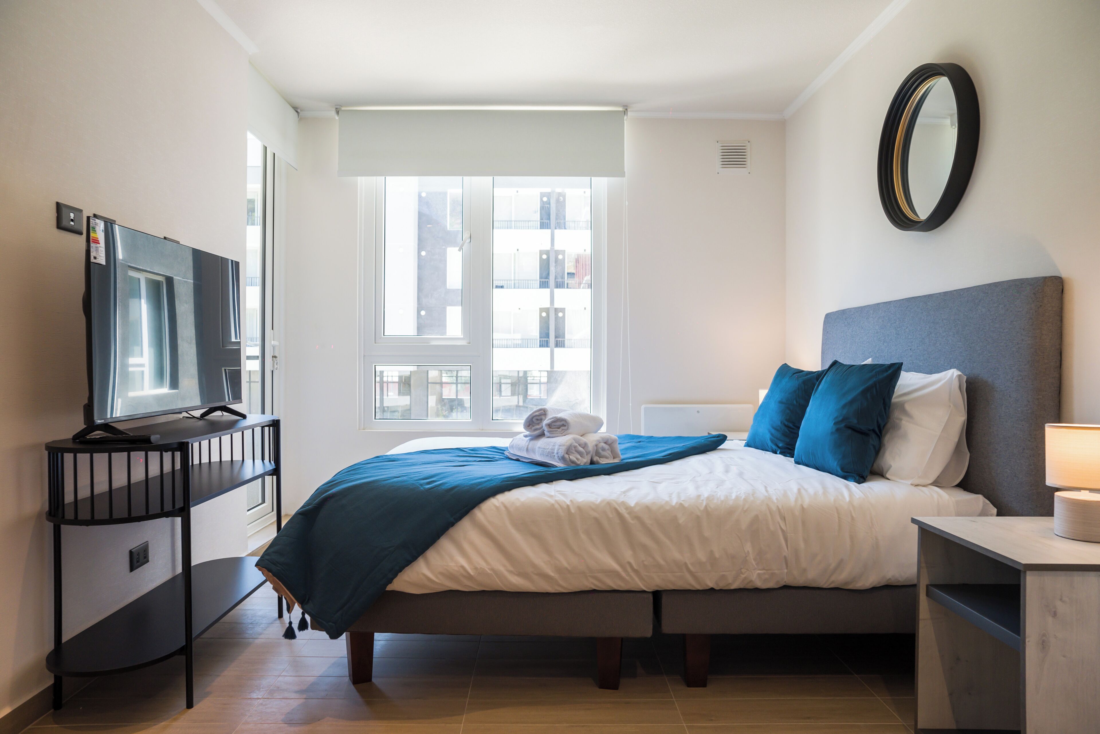 A hotel room with a bed, a television on a stand, a window with a view of buildings, and a bedside table with a lamp.