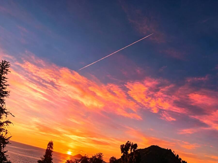 Un atardecer con cielo despejado, una estela visible y siluetas de árboles y una montaña.