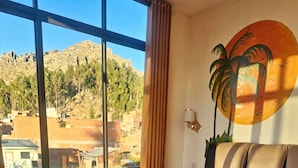 A hotel room with a bed, a window with a view of a mountainous landscape, and a wall with a palm tree mural.