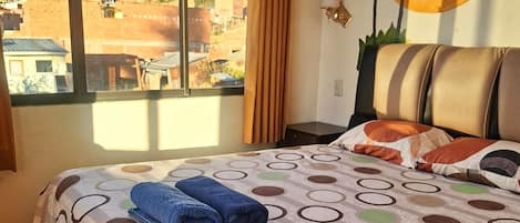 A hotel room with a bed, a window with a view of a mountainous landscape, and a wall with a palm tree mural.
