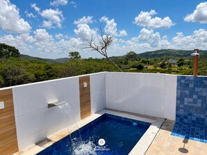 A swimming pool with a waterfall feature, surrounded by white and blue tiles, set against a backdrop of a clear sky and a hilly landscape.