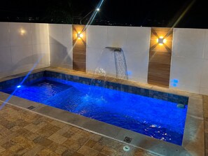 A rectangular swimming pool with a waterfall feature, surrounded by tiled walls and illuminated by wall-mounted lights.