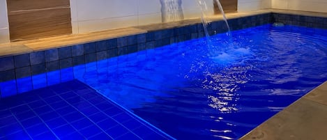 A swimming pool with a waterfall feature, surrounded by white tiled walls and wooden accents.