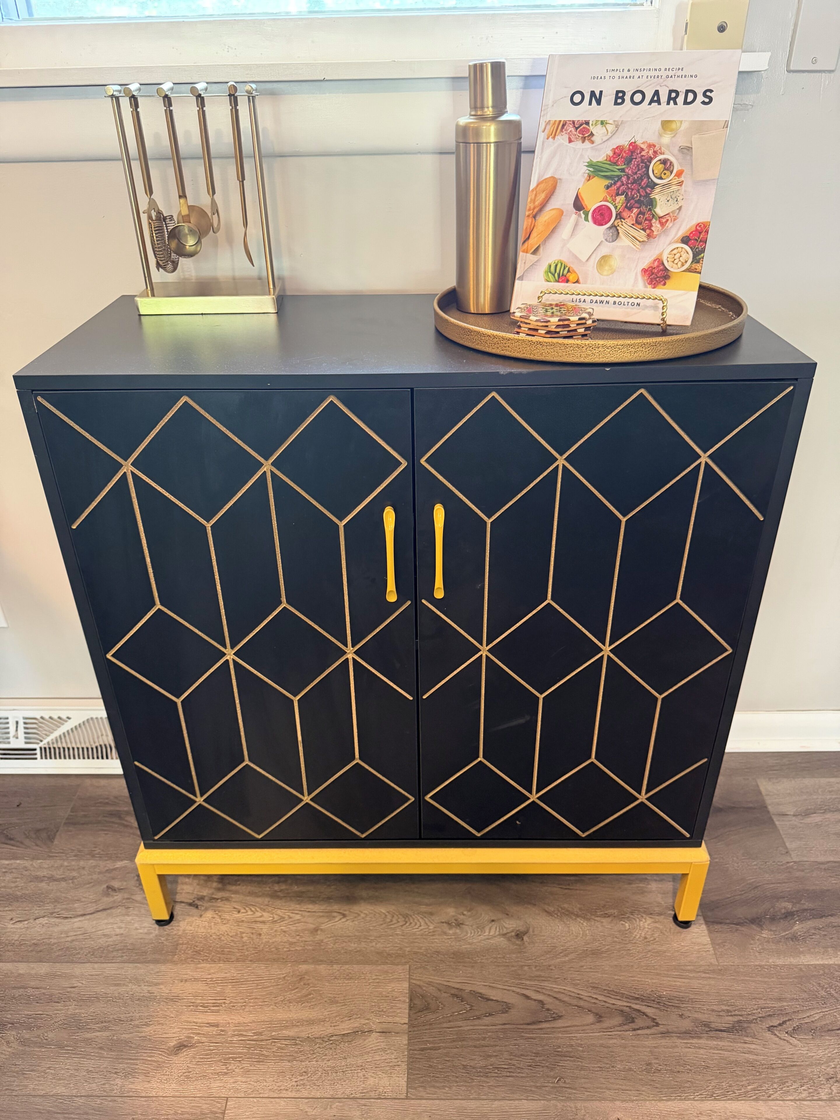 A dark blue cabinet with geometric gold patterns, yellow handles, and a tray with a cookbook on top.