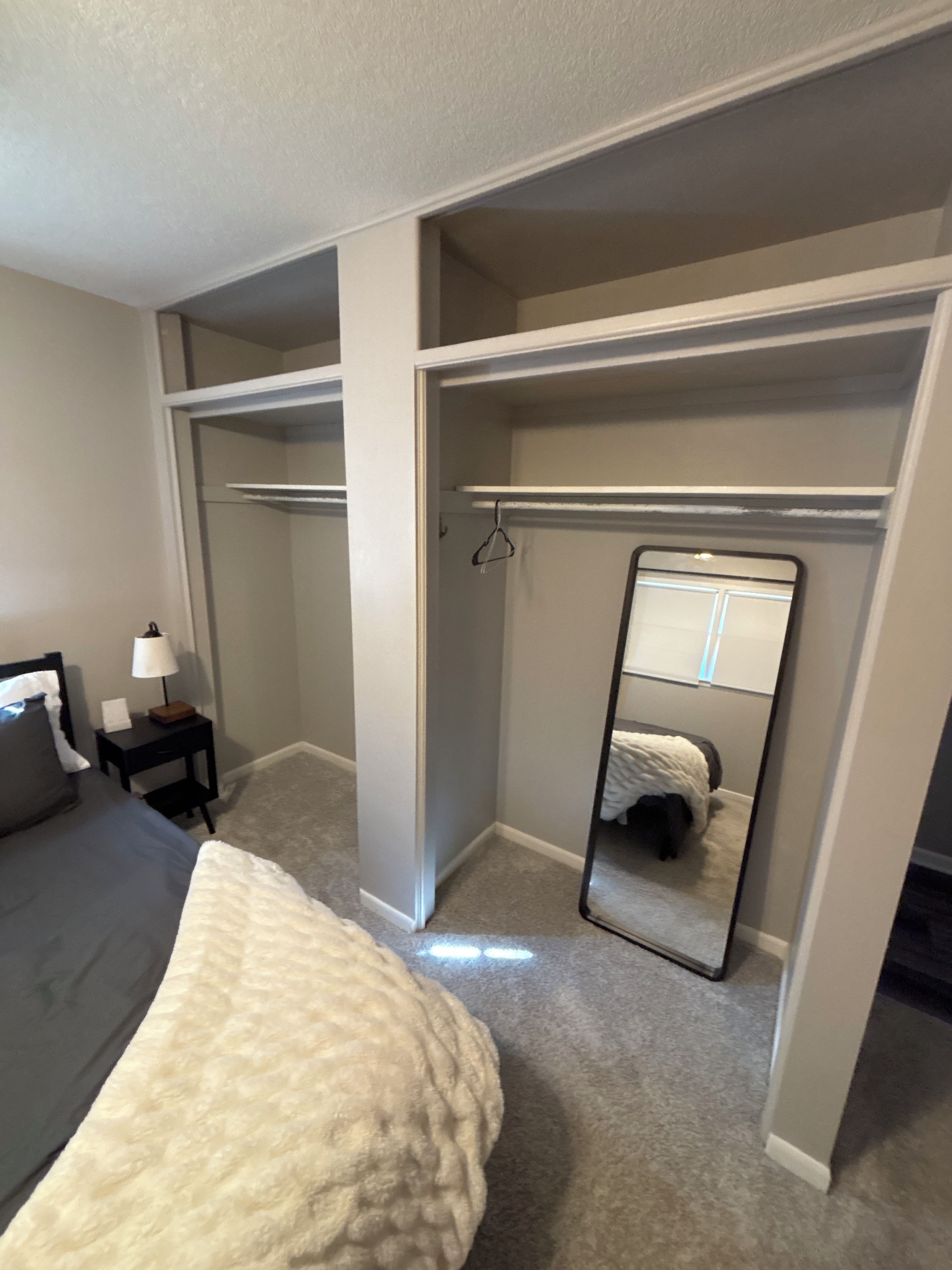 A bedroom with a bed, a nightstand, a bedside table, a closet, and a full-length mirror.