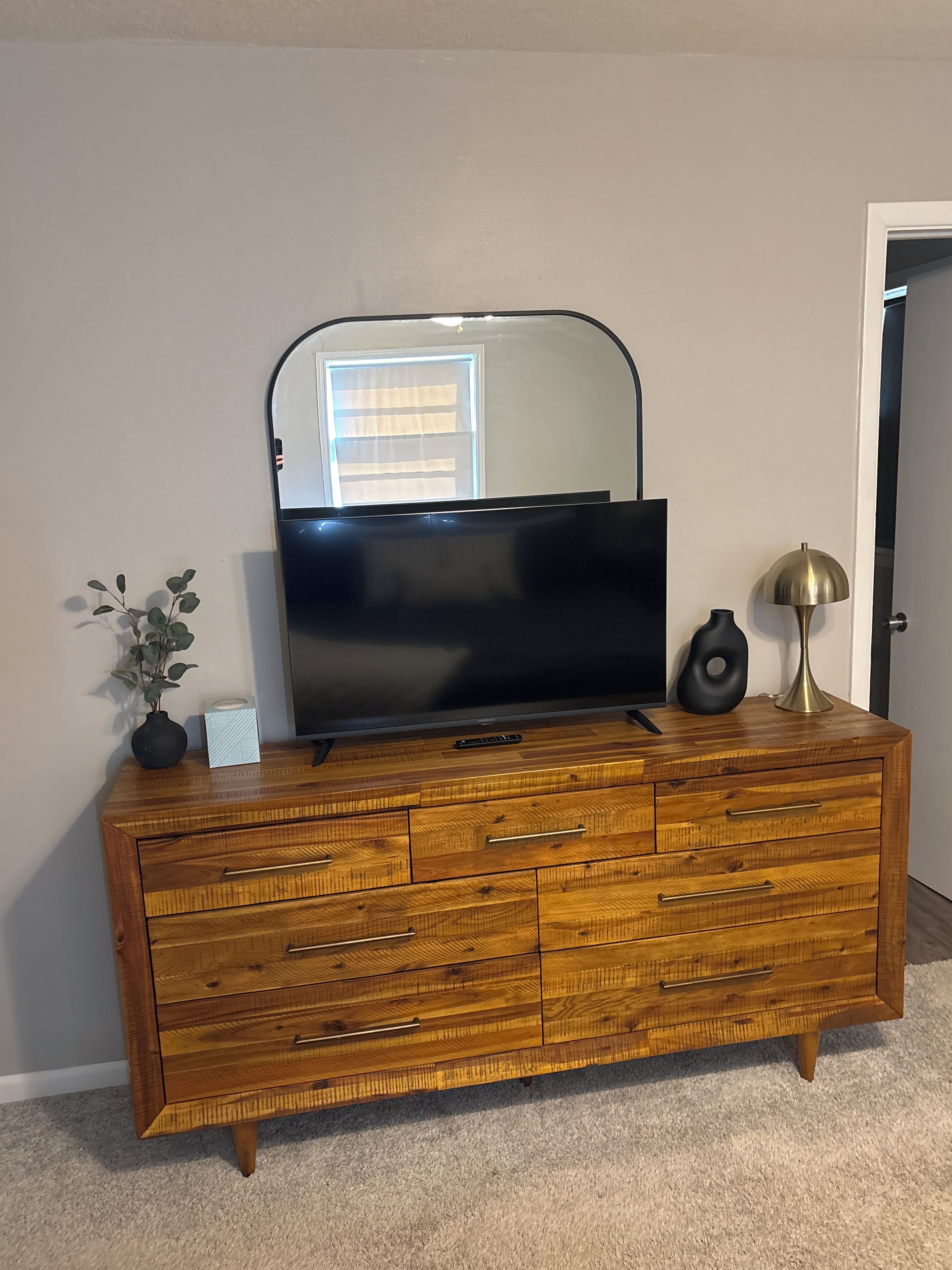 A wooden dresser with a flat-screen TV, a mirror, a vase, and a lamp.