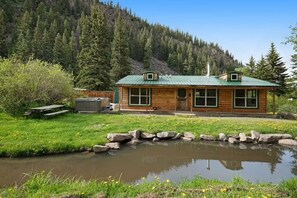 A cabin with a green roof, a pond in front, and a forested hillside in the background. - Ponderosa | Creekside Cabin Near Wolf Creek (South Fork)