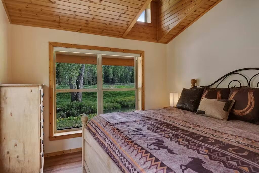 A bedroom with a large window, a bed with patterned bedding, and a wooden ceiling.