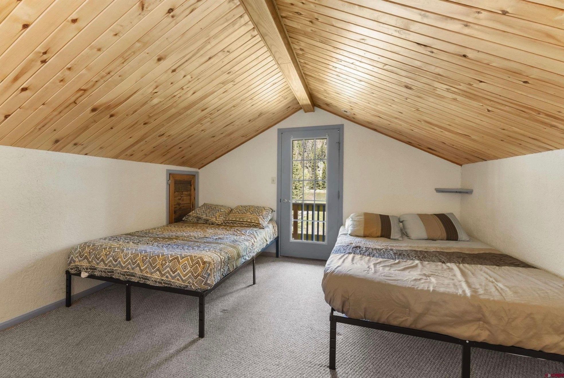 A small attic bedroom with two single beds, a wooden sloped ceiling, and a door leading outside.