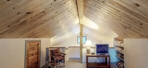 Ein Zimmer mit schrägem Dach, einer Holztür, einem gemusterten Sessel, einem Tisch mit Lampe und einem Fernseher.