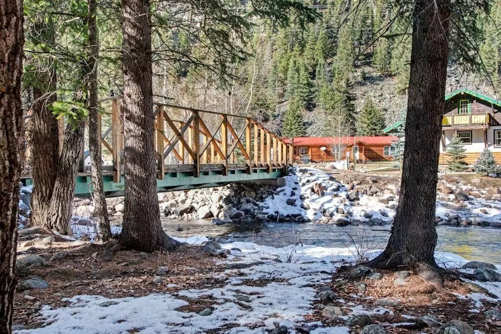 A wooden bridge over a river, surrounded by trees and a cabin with a red roof.
