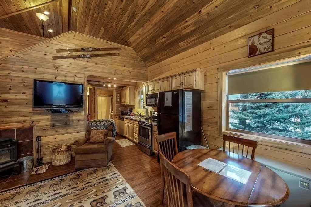 Ein Holzblockinnenraum mit Esstisch, Stühlen, Fernseher, Küchenzeile und einem Fenster mit Blick auf Bäume.