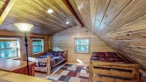 Mökki, jossa on kalteva katto, puuseinät, ruutukuviollinen lattia ja kaksi sänkyä kuviollisilla vuodevaatteilla.