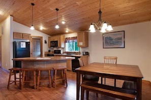 A kitchen with wooden cabinets, a refrigerator, a microwave, and a dining area with a wooden table and chairs. - Sequoia Cabin | Waterfront Escape with Private Hot (South Fork)