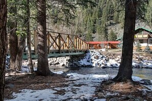 Un pont en bois enjambant une rivière, entouré d’arbres et bordé d’une cabane au toit rouge.