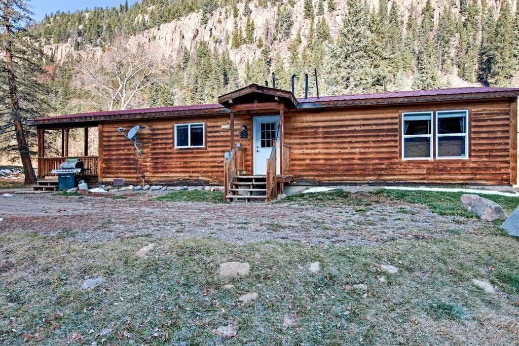 A wooden cabin with a front porch, a chimney, and a view of a forested mountain range.