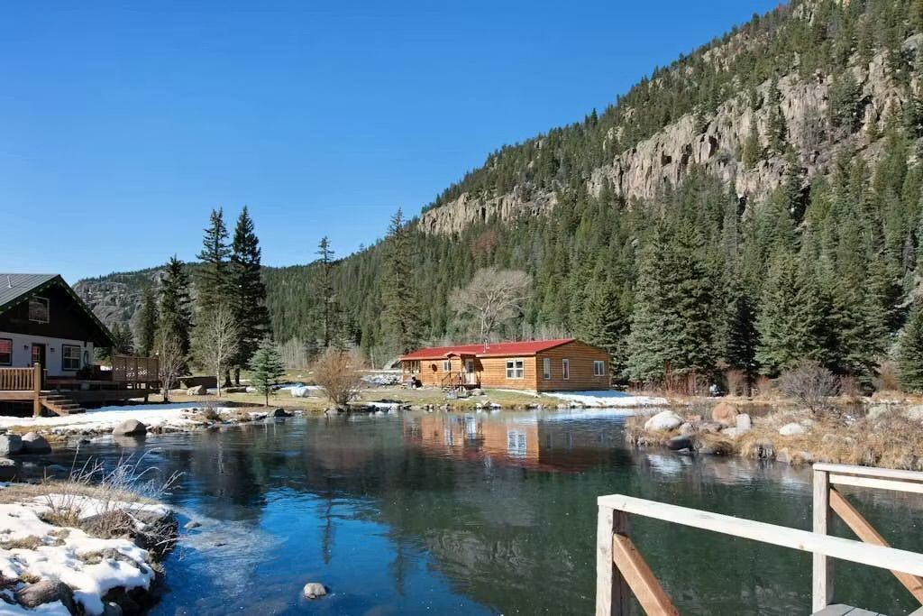Une cabane avec un toit rouge, située près d’une rivière et entourée de montagnes boisées.