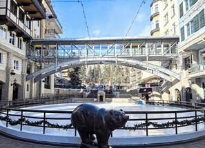 Ein öffentlicher Platz mit einem zentralen Brunnen, umgeben von Steingebäuden mit Balkonen, unter einer Fußgängerbrücke.