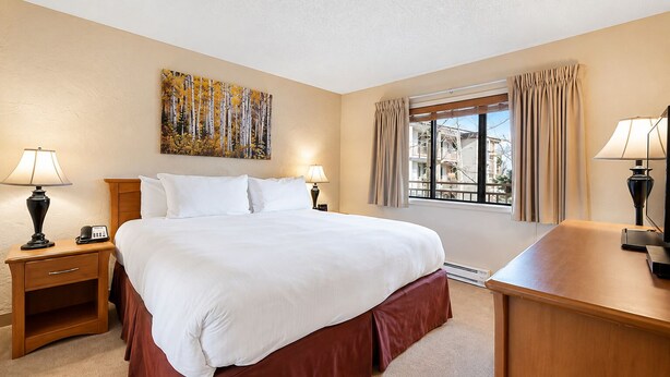 A hotel room with a large bed, bedside lamps, a desk, a window with curtains, and a painting on the wall. - 1 BR+ sofabed & fireplace, walk to Village, Shuttle, Hot Tubs, 3/21-27/2027 ONLY (Vail)