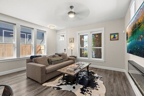 A living room with a sofa, coffee table, and a cowhide rug. There are windows, a ceiling fan, and artwork on the walls.
