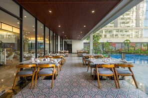 En restaurang med utsikt över poolen, med en lång matsal med bord och stolar, ett mönstrat golv och ett träpanelat tak.