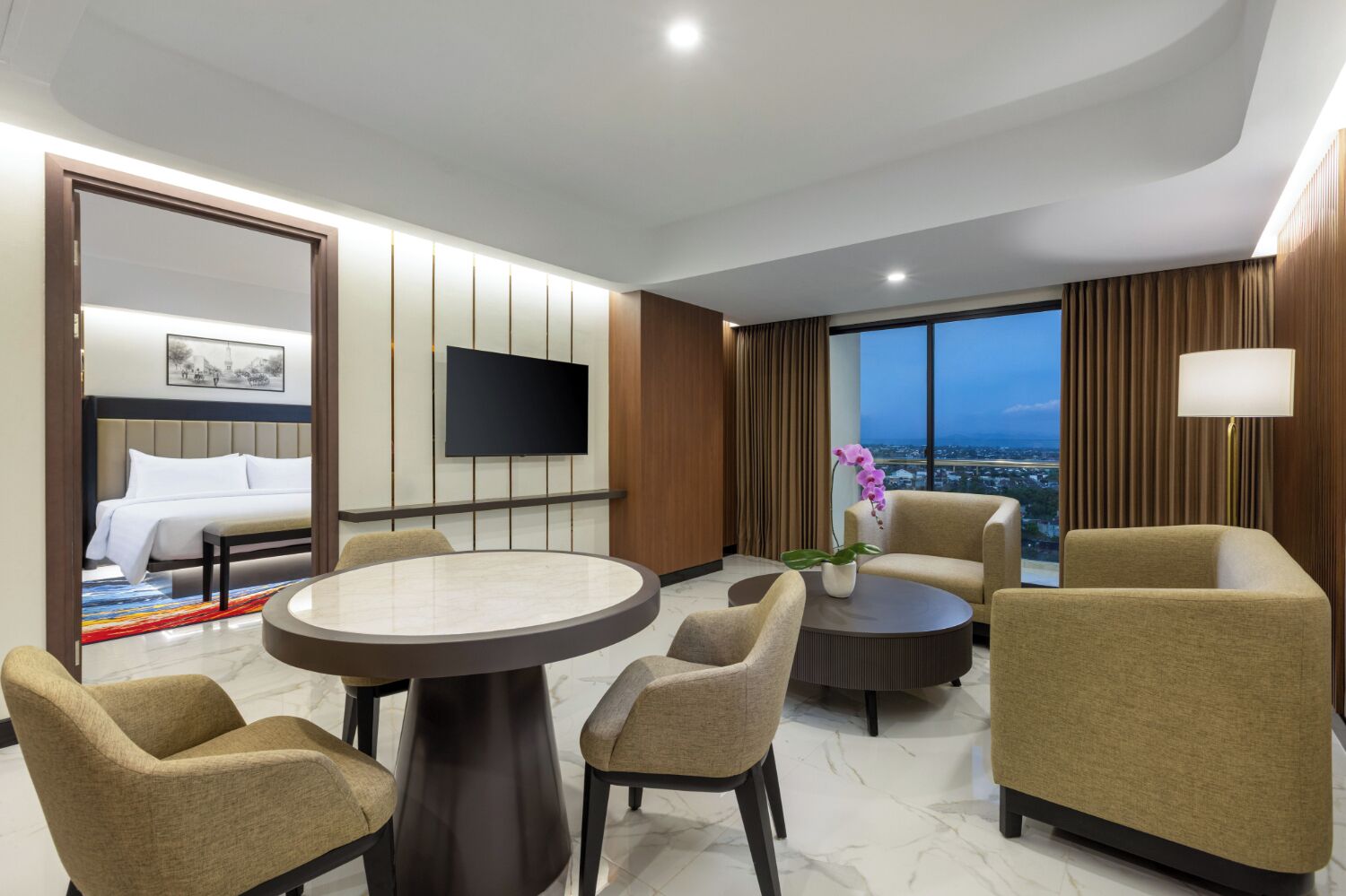 A hotel room with a bed, a TV, a dining table, and a view of the city.