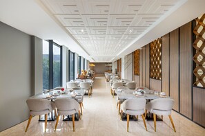 A long dining area with tables and chairs, a patterned ceiling, and large windows. - The Royal Alana Yogyakarta Hotel & Convention Center (Ngaglik)