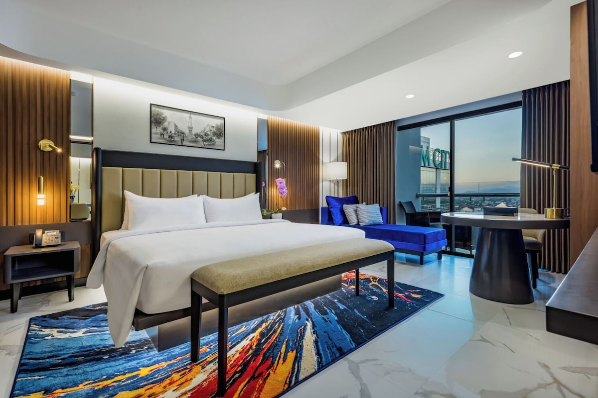 Quarto de hotel moderno com uma cama grande, uma escrivaninha, uma cadeira e vista para a cidade.