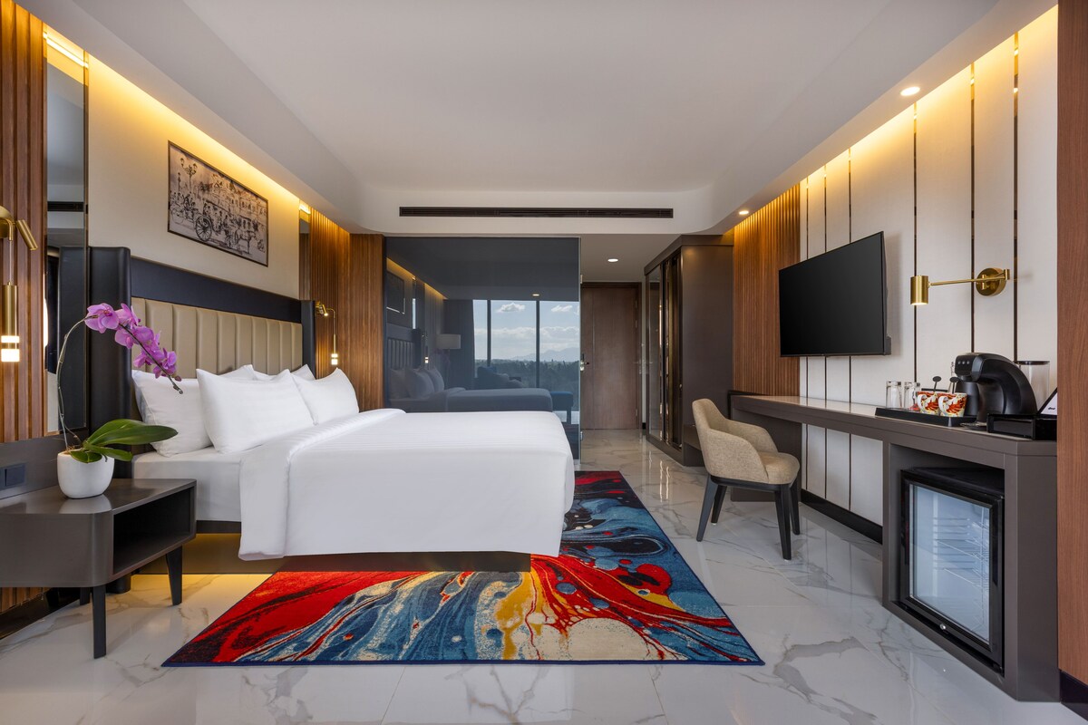 A modern hotel room with a large bed, a desk with a flat-screen TV, and a view of the city.