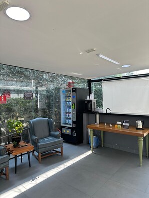 A modern room with a seating area, a vending machine, a coffee station, and a view through large windows. - ADD HOTÉIS MORUMBI (São Paulo)