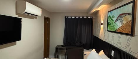 A hotel room with a bed, a flat-screen TV mounted on the wall, an air conditioning unit, a desk with a chair, and a painting on the wall.