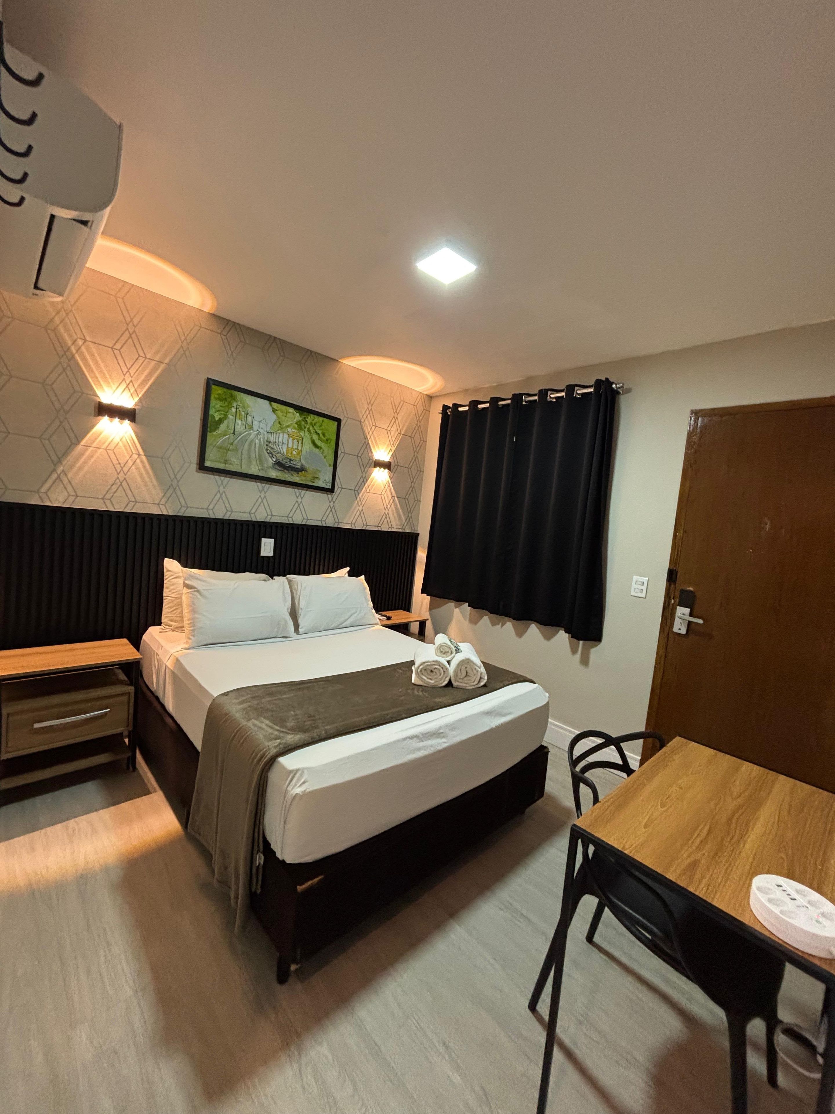 A hotel room with a bed, bedside table, a desk, and a chair. There is a painting on the wall and a door to the right.