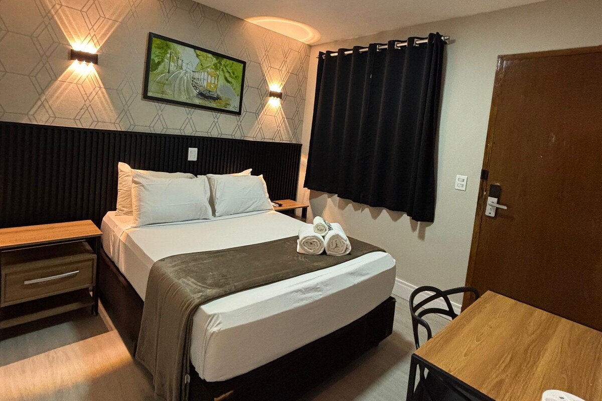 A hotel room with a bed, bedside table, a desk, and a chair. There is a painting on the wall and a door to the right.
