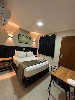 A hotel room with a bed, bedside table, a desk, and a chair. There is a painting on the wall and a door to the right.