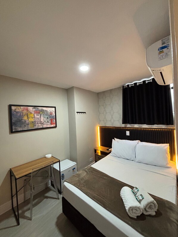 Habitación de hotel con cama, escritorio, silla, aire acondicionado de pared y un cuadro.