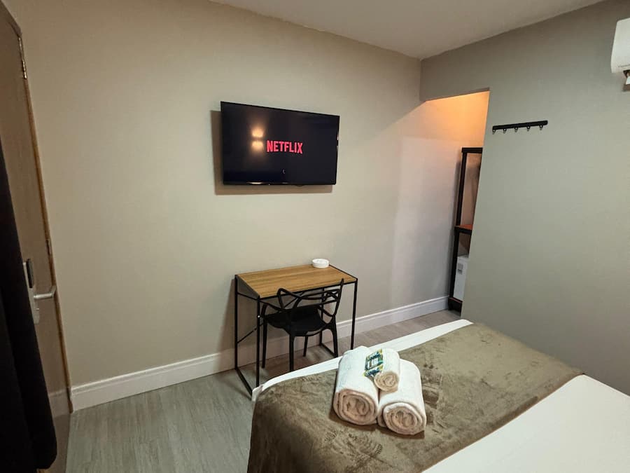 Quarto de hotel com uma cama, uma escrivaninha com cadeira, uma televisão na parede e uma mesinha com uma tigela.