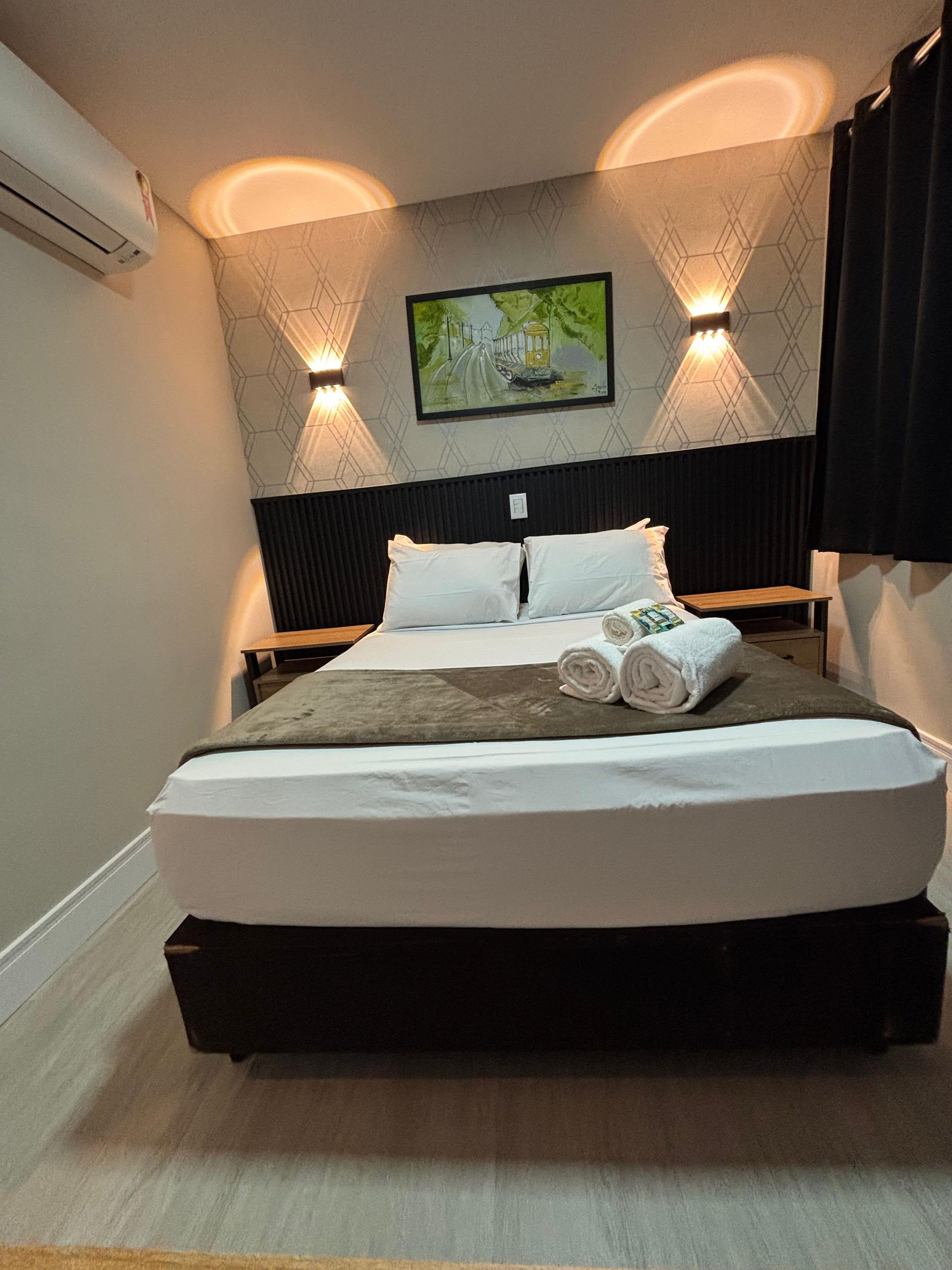 Habitación de hotel con una cama grande, dos mesitas de noche, aire acondicionado de pared y un cuadro sobre la cama.