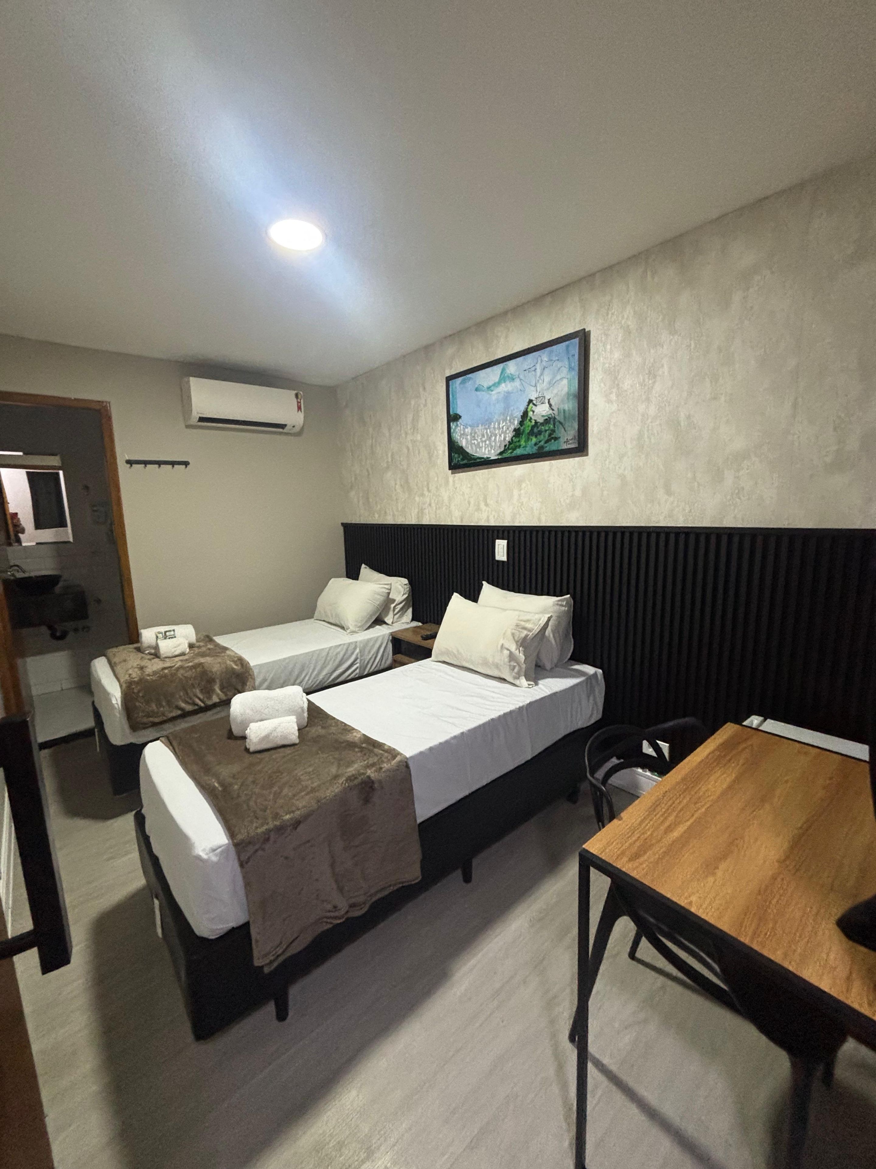 Ein Hotelzimmer mit zwei Betten, einem Holztisch, einem Bild an der Wand und einer Klimaanlage.