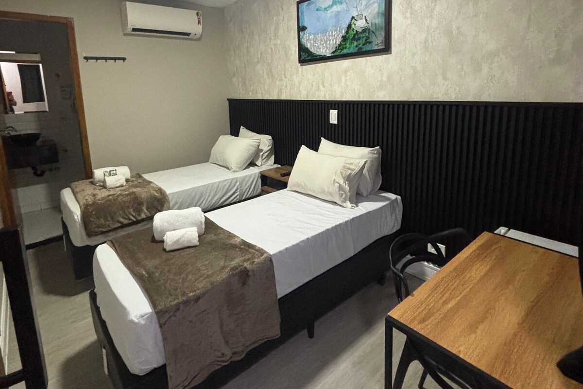 A hotel room with two beds, a wooden table, a painting on the wall, and an air conditioning unit.