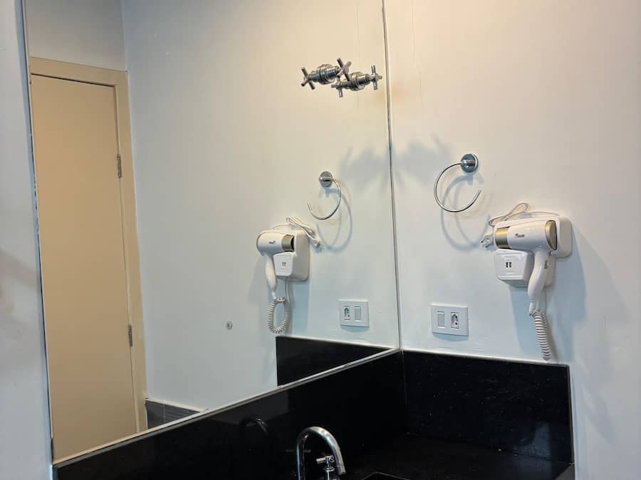 Casa de banho com lavatório preto, dois secadores de cabelo fixados na parede e espelho sobre o lavatório.