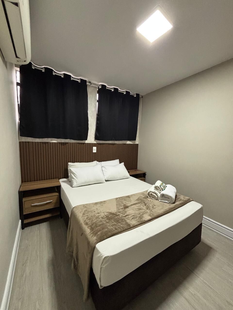 Quarto de hotel com cama, mesas de cabeceira, cortinas e luminária no teto.